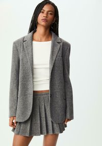 Blazer grigio in misto lana su misura con rever, abbinato a una gonna mini plissettata coordinata. Sotto, un top bianco aderente.