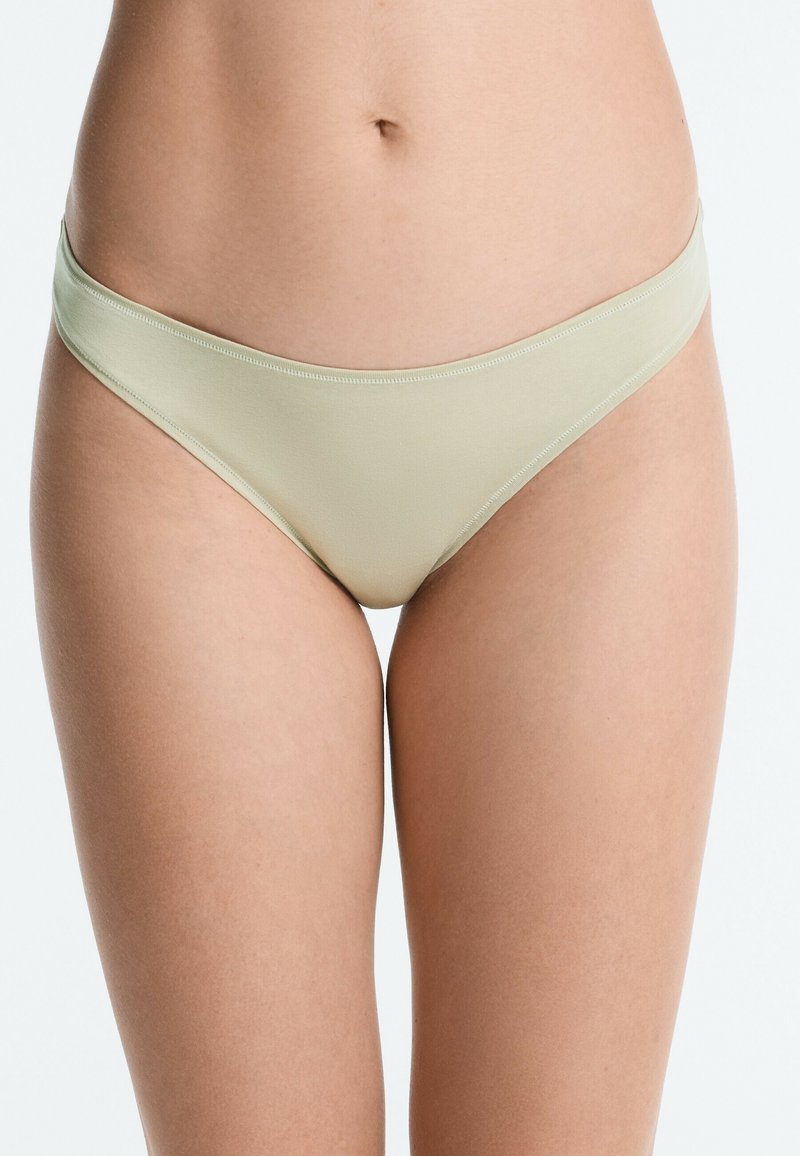 Taille et parties supérieures des cuisses féminines portant un sous-vêtement style bikini beige clair, taille basse et sans couture, sur fond uni.