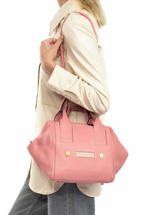 Vrouw met een beige jas en grijze broek die een roze gestructureerde handtas over haar schouder draagt tegen een witte achtergrond.