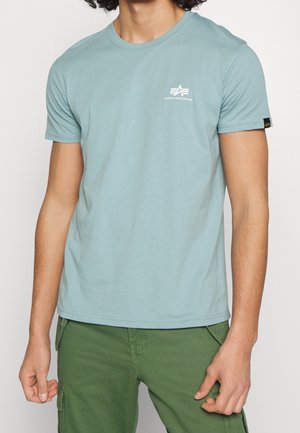 Homme portant un t-shirt bleu clair Alpha Industries et un pantalon cargo vert, montré du cou aux hanches sur un fond blanc uni.