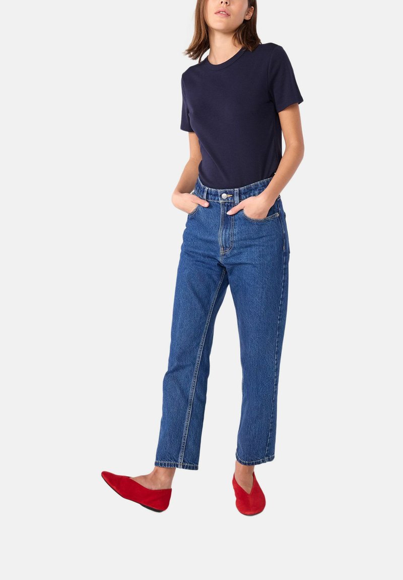 Donkerblauw t-shirt gecombineerd met lichtblauwe denim jeans met hoge taille. Model draagt rode ballerina's, met de handen in de zakken, en toont zo een casual look.