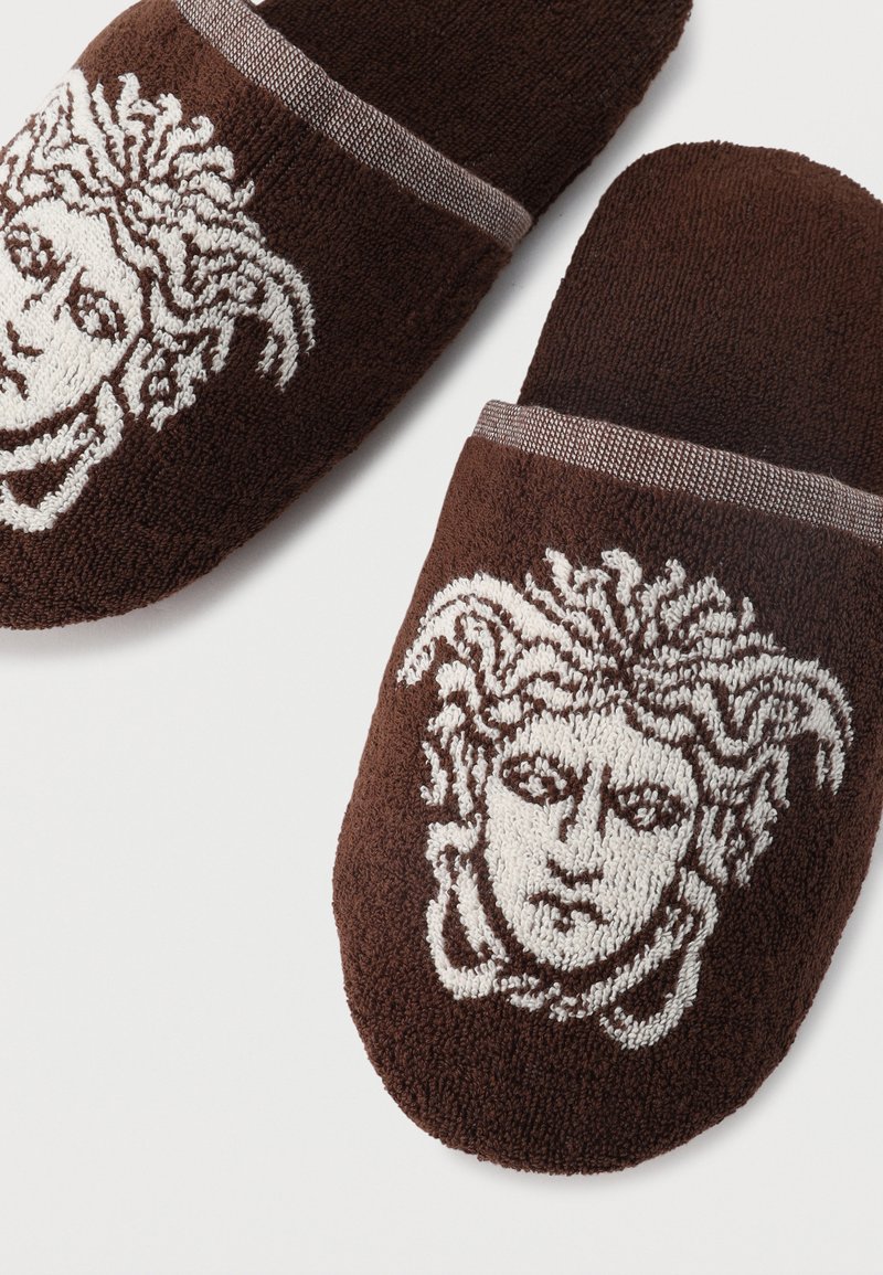 Pantofole in spugna marrone con ricamo bianco della testa di Medusa sulla parte superiore, caratterizzate da tessuto morbido e design a calzata.