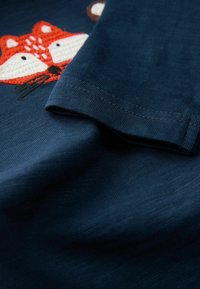 Mornarsko plavo tkanje s rebrastom teksturom sadrži izvezenu dizajn lisice u narančastoj i bijeloj boji, smješten ispred.