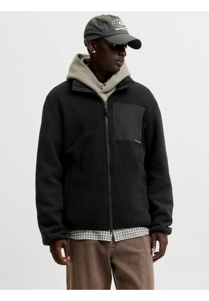 Homme portant une veste polaire noire zippée par-dessus un sweat à capuche beige, une chemise à carreaux, un pantalon marron et une casquette grise, regardant sur le côté.