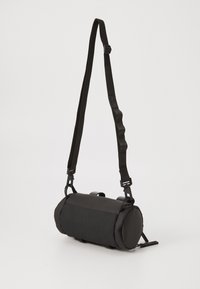Borsa a tracolla cilindrica nera con tracolla regolabile e manico di trasporto piccolo su uno sfondo bianco semplice.
