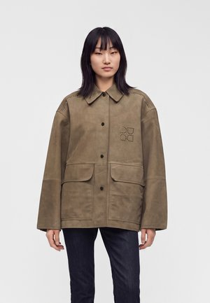 Personne aux cheveux noirs et raides porte une veste oversize vert olive avec poches avant et fermeture boutonnée sur un pantalon sombre, sur un fond uni.
