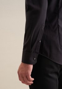 Braccio sinistro di un uomo in camicia nera a maniche lunghe con polsino abbottonato, mano rilassata e pantaloni neri su sfondo neutro.