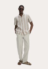 Man in een beige en wit gestreept shirt met korte mouwen, lichte beige broek, beige sandalen en een donkere zonnebril, staande met de handen in de zakken.