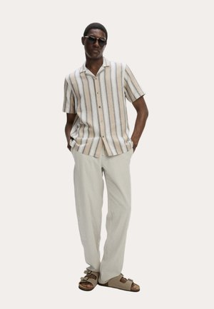 Hombre con camisa de manga corta a rayas beige y blanca, pantalones beige claros, sandalias beige y gafas de sol oscuras, de pie con las manos en los bolsillos.