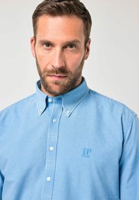 Mies, jolla on lyhyet ruskeat hiukset ja parta, pukeutuneena vaaleansiniseen, napitettuun paitaan, jossa on rintataskussa kirjailtu "JP 1850", yksiväristä taustaa vasten.