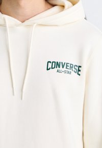 Sweat à capuche couleur crème en tissu doux, avec un logo vert "CONVERSE ALL STAR" sur la poitrine et des cordons de serrage sur la capuche.