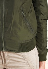 Olivově zelená bomber bunda s kapsou na zip, žebrovanými manžetami a hladkým, lesklým povrchem. Zapínání na zip se táhne po přední části.