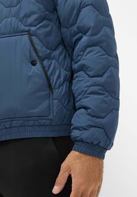 Blå kviltet pufferjakke med en struktureret overflade, der har en lynlåslomme, elastiske mansjetter og en tætsiddende kant for varme og komfort.