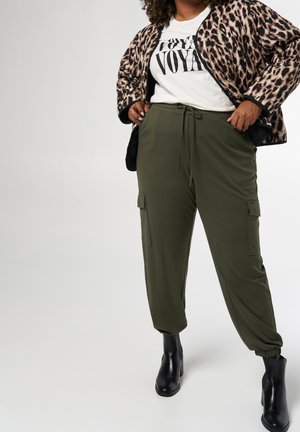 Personne aux formes portant une veste imprimée léopard, un t-shirt graphique blanc, un pantalon cargo vert olive et des bottines noires, debout les mains dans les poches.