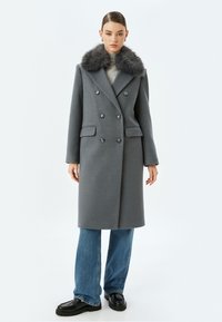 Abrigo de lana gris con un diseño de doble botonadura, cuello de piel sintética y dos bolsillos, llevado sobre jeans de pierna ancha y mocasines negros.