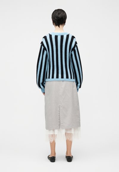 Μπλε και μαύρο ριγέ πουλόβερ με χαλαρούς ώμους, σε συνδυασμό με γκρι φούστα που έχει σκίσιμο πίσω πάνω από λευκή δαντέλα. Μαύρα επίπεδα παπούτσια.