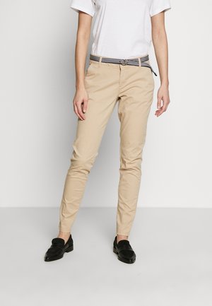 Person iført beige slim-fit bukser, sorte loafers, sort-hvid stribet bælte og en enkel hvid kortærmet skjorte, stående.