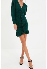 Robe verte à manches longues avec un ourlet volanté. Dotée de fronces sur les côtés et d'un décolleté en V plongeant. Associée à des talons noirs à lanières.