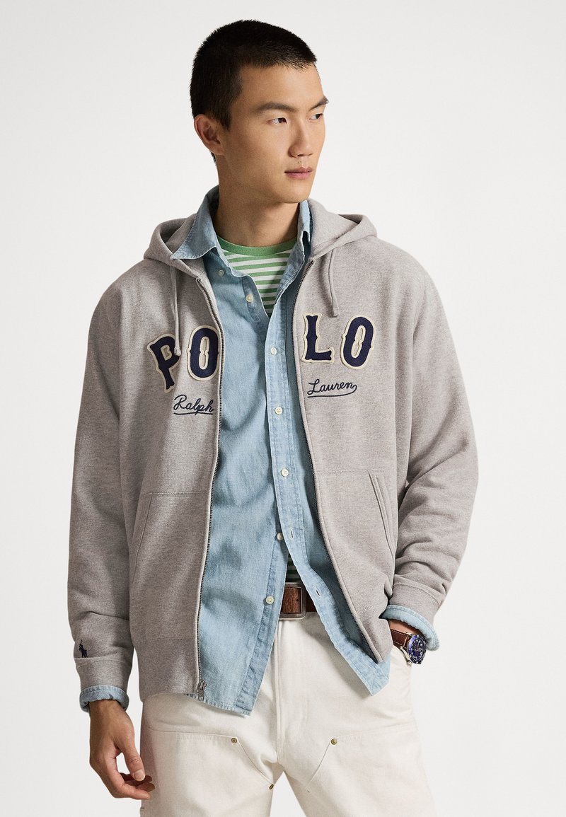 Άνδρας που φοράει γκρι φούτερ με φερμουάρ και το �γράμμα «POLO», μπλε τζιν πουκάμισο, πράσινη ριγέ μπλούζα, λευκό παντελόνι και ρολόι χειρός, κοιτάζοντας προς τα δεξιά.