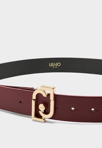 Cintura in pelle bordeaux con una texture liscia, una fibbia dorata dalla forma unica e un interno nero con logo impresso.