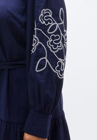 Φόρεμα σε ναυτικό  μπλε με μακριά μανίκια, λευκά κεντήματα με λουλούδια, άνετη γραμμή και βολάν στο τελείωμα. Υφή υφάσματος.