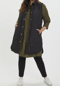 Schwarze gesteppte Weste mit hohem Kragen, Seitentaschen und Druckknopfverschluss, darüber ein olivgrünes Hemdkleid und schwarze Hose. Weiße Sneakers.