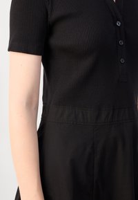Black dress with short ribbed sleeves, buttoned placket, and a smooth lower fabric. Features a fitted bodice transitioning to a gently flared skirt.