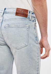 Svijetloplave traperice imaju stražnji džep, glatku teksturu i smeđu kožnu zakrpu s utisnutim logotipom na struku.