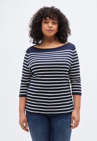 Μπλε ναυτικό και λευκό ριγέ μακρυμάνικο πουκάμισο με λαιμόκοψη σκάφος, με απαλή υφή και μανίκια μέτριου μήκους. Συνδυασμένο με μπλε τζιν παντελόνι.