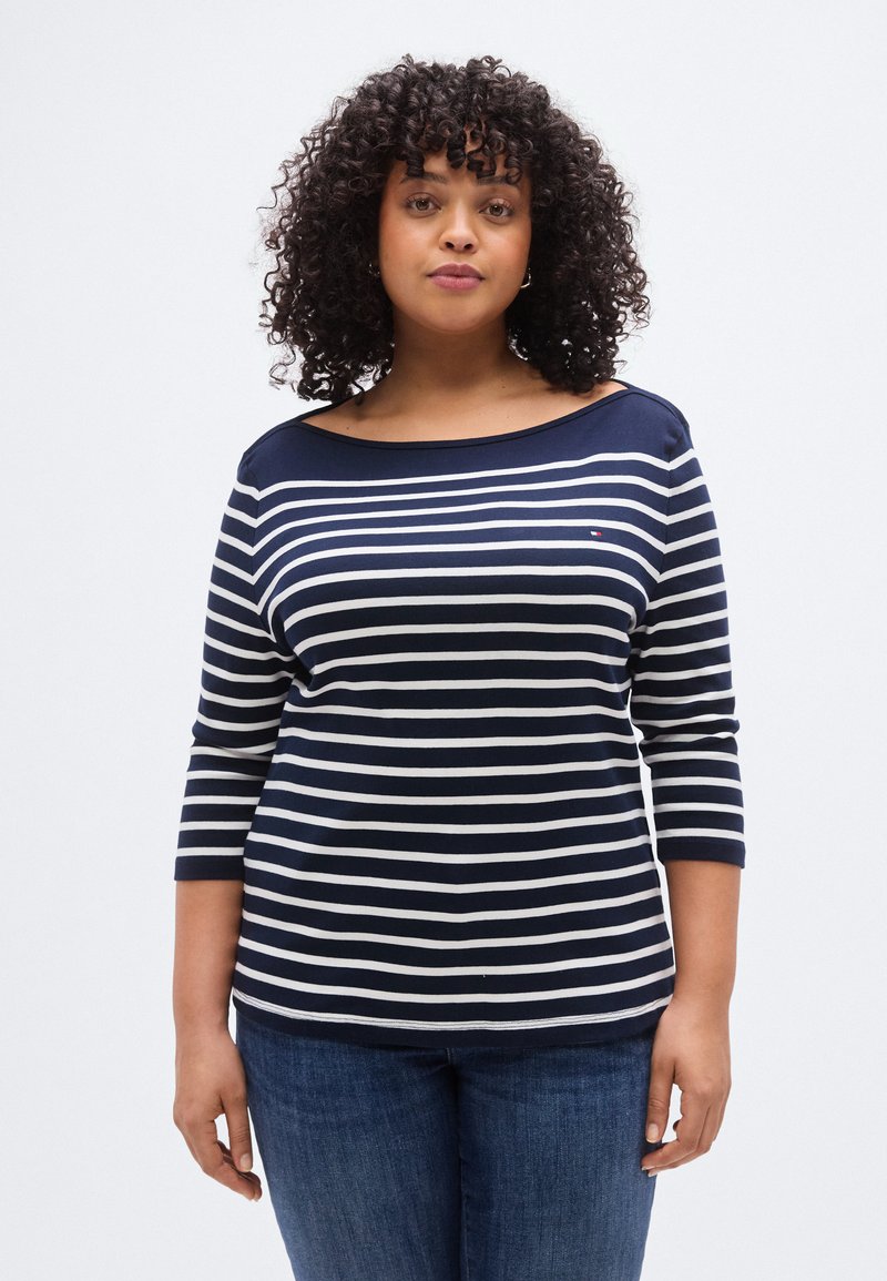 Μπλε ναυτικό και λευκό ριγέ μακρυμάνικο πουκάμισο με λαιμόκοψη σκάφος, με απαλή υφή και μανίκια μέτριου μήκους. Συνδυασμένο με μπλε τζιν παντελόνι.