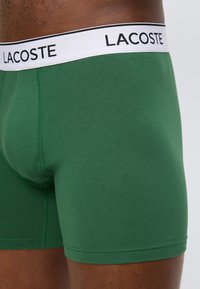Πράσινα ανδρικά μπόξερ με λευκή μέση στη μέση που φέρει τη σήμανση "LACOSTE", φορεμένα από ένα άτομο πάνω σε ουδέτερο φόντο.