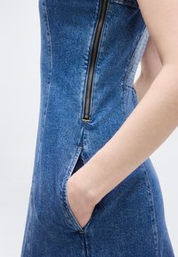 Άτομο που φοράει μπλε τζιν φόρεμα με φερμουάρ στο πλάι και το χέρι στην τσέπη, απεικονιζόμενο από τη μέση έως το μέσο του μηρού με φόντο απλό.