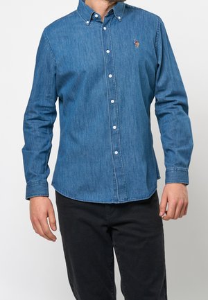 Mand iført langærmet blå denim skjorte med krave og broderet logo på brystet, kombineret med sorte bukser.