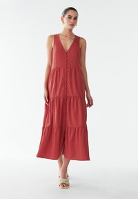 Maxikleidi tellises punases kangas, millel on sügav V-kaelus, ees nupud ja kihiline disain. Sile tekstuur ja keskmise pikkusega alumine serv.