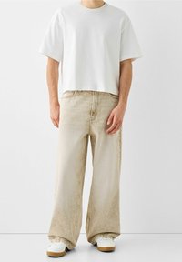Vit kortärmad t-shirt, beige vida jeans med blekt mönster och vita sneakers med gummisulor. En enkel, avslappnad outfit.