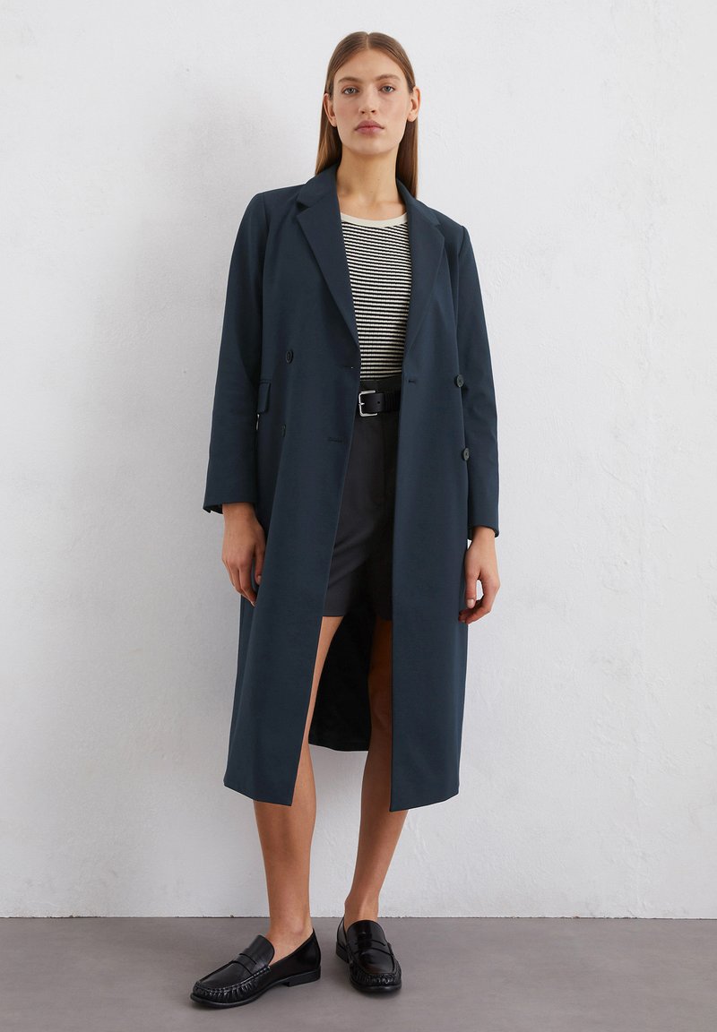 Langer dunkeltürkiser Mantel mit doppelreihigem Design, Spitzenlazetten und seitlichen Schlitzen, kombiniert mit einem gestreiften Oberteil, Shorts und schwarzen Loafers.