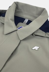 Jasnoszara kurtka z zagiętym kołnierzem, z białym logo na lewej piersi i kontrastowym granatowym wykończeniem widocznym w środku.