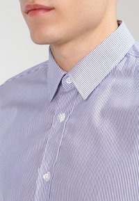 Randig skjortklänning med knäppbar krage, med smala lila och vita ränder, samt en mjuk bomullstextur. Två framknappar synliga.