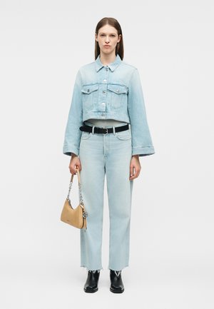 Sieviete valkā gaiši zilu īsu džinsa jaku un džinsus, melnu jostu, melnus zābakus, tur rokā mazu bēša krāsas somiņu ar ķēdes siksnu.