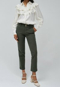 Bílá blůza s vrstvenými volány, spárovaná s olivově zelenými úzkými kalhotami. Modelka má na sobě béžové boty na blokovém podpatku. Jednoduchý, elegantní design.