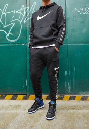 Osoba ubrana w czarną bluzę z kapturem Nike i joggery cargo z białymi logo Swoosh, stoi przy zielonej metalowej ścianie pokrytej graffiti.
