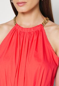 Gros plan sur une femme portant un haut en corail à col halter avec une sangle en chaîne dorée arborant des lettres en métal audacieuses.