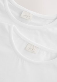 Deux t-shirts en coton blanc empilés, avec des encolures rondes. Les étiquettes indiquent la marque "NEXT" et le tissu fabriqué au Sri Lanka. Texture lisse.