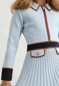Svetlomodré šaty s dlhými rukávmi, predným zipsom, hnedými akcentmi na golieri a páse a skladanou sukňou s detailom zlatého gombíka.