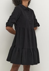 Schwarzes Baumwollkleid mit einem Knopfkragen, langen Ärmeln, die bis zum Ellbogen hochgekrempelt sind, und einem mehrstufigen Volant-Saum; glatte Textur und entspannte Passform.