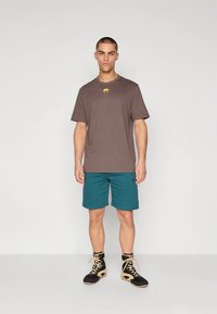 Ruda medvilninė marškinėliai su geltonu logotipu, derinami su tamsiai žaliais šortais ir juodais batais. Paprastas dizainas, laisvas prigludimas ir kontrastingos spalvos.