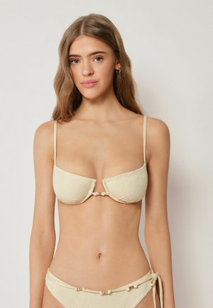 Kvinde med langt, bølget brunt hår iført en cremefarvet tekstureret bikini med tynde stropper og små sløjfedetaljer, stående mod en ensfarvet baggrund.