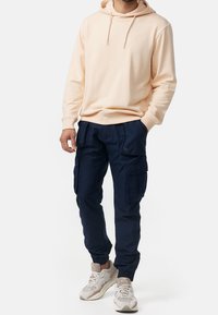 Licht perzikkleurige hoodie met een trekkoord en een logodetail, gecombineerd met marineblauwe cargobroek met meerdere zakken en elastische boorden.
