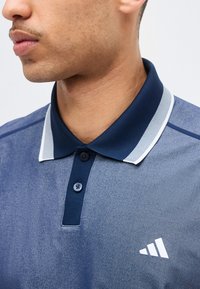 Άνδρας που φοράει μπλε πόλο πουκάμισο με ναυτικό γιακά και δύο κουμπιά, λευκή ριγέ λεπτομέρεια στον γιακά και λευκό λογότυπο Adidas στο στήθος.