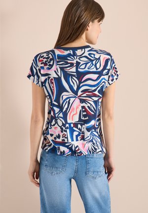 Femme portant une chemise à manches courtes à motif abstrait multicolore et un jean en denim bleu clair, tournée de dos face à la caméra sur un fond beige.
